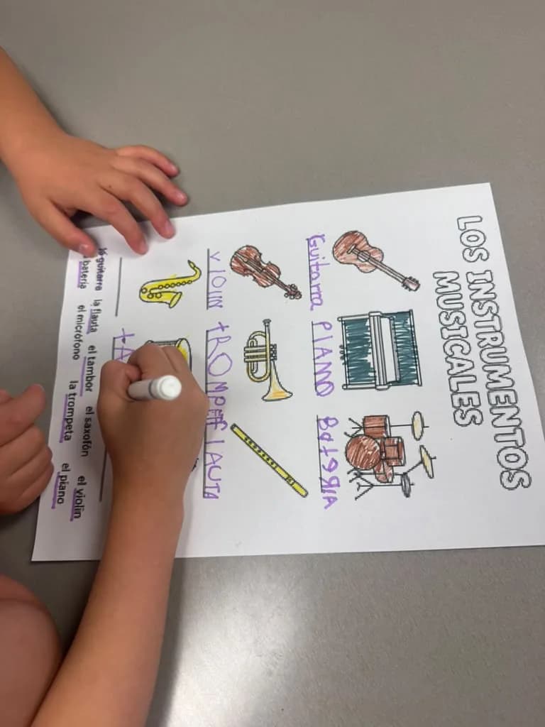 A student's hands writing the Spanish names of instruments on a worksheet with a marker