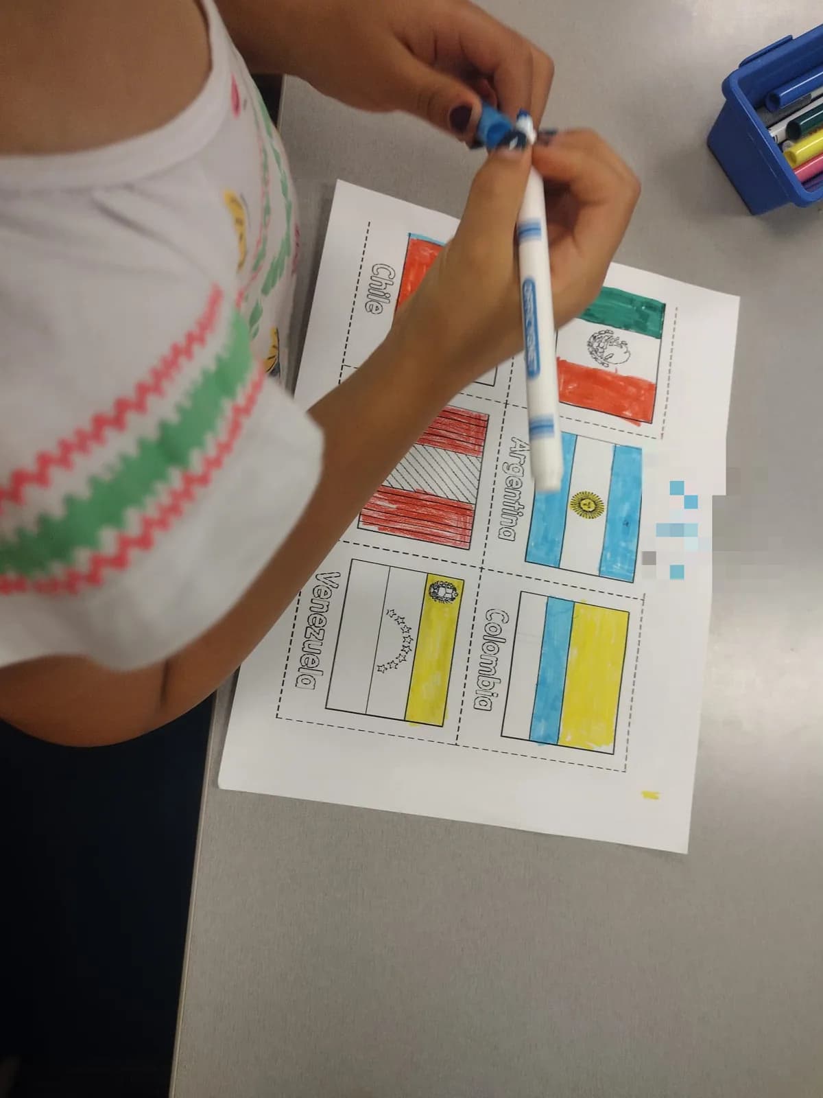 A student reaching across a table with a marker to finish colouring a flags worksheet