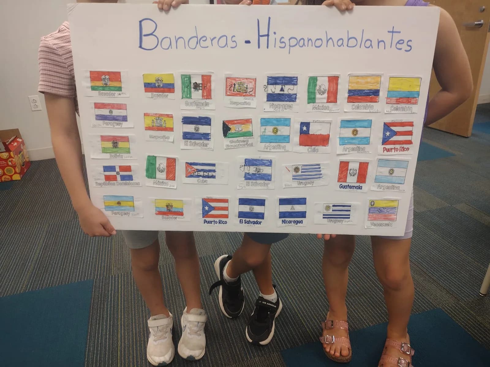 Three students holding up a large poster covered in hand-drawn flags of Spanish-speaking countries